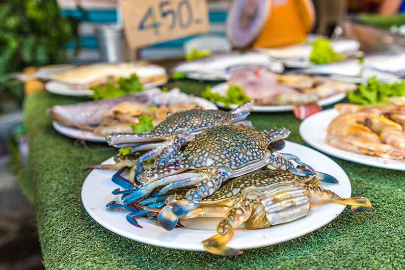 Frisches Seafood auf einem Marktstand mit grünem Untergrund. Im Vordergrund sind mehrere blau-gesprenkelte Krabben kunstvoll auf einem weißen Teller angerichtet. Im leicht unscharfen Hintergrund stehen weitere weiße Teller mit frischem Fisch, Garnelen und Salatblättern sowie ein Pappschild mit der handgeschriebenen Preisangabe 450. Koh Lipe Thailand Seafood