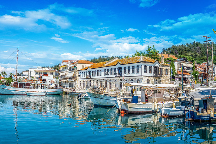 Ein kleiner Hafen mit mehreren Booten liegt vor einer Uferpromenade mit traditionellen Gebäuden und Cafés. Das ruhige Wasser spiegelt die Boote und die Architektur wider, während im Hintergrund Bäume und weitere Häuser zu sehen sind. Thassos Limenas