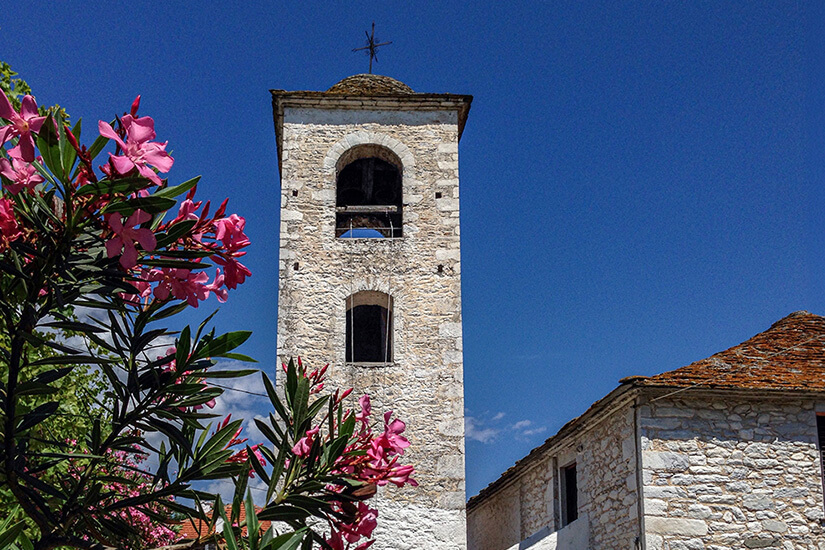 Ein schlichter Kirchturm aus hellem Naturstein ragt vor einem tiefblauen Himmel empor. Im Vordergrund setzen blühende rosa Sträucher einen farbigen Akzent, während rechts ein weiteres traditionelles Steingebäude zu sehen ist. Thassos Theologos