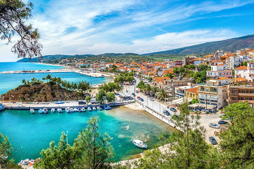 Eine malerische Küstenstadt erstreckt sich entlang einer türkisfarbenen Bucht mit kleinen Booten und einem geschützten Hafen. Entlang der Uferstraße reihen sich Wohnhäuser, Hotels und Restaurants mit Blick auf das klare Wasser. Im Hintergrund steigen bewaldete Hügel sanft an und rahmen die Küstenlandschaft ein. Thassos Limenaria