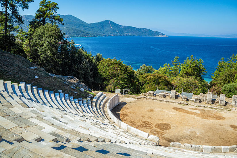 Ein antikes Freilufttheater mit halbkreisförmig angeordneten Steinstufen liegt an einem Hang über dem Meer. Hinter der Bühne erstrecken sich dichte Wälder, während die Küste und das tiefblaue Wasser einen eindrucksvollen Hintergrund bilden. Griechenland Theater