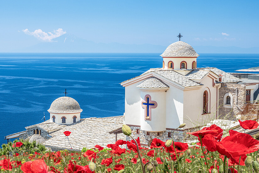 Eine helle Kirche oder Klosteranlage mit kuppelförmigen Dächern liegt auf einer Anhöhe über dem tiefblauen Meer. Im Vordergrund leuchten rote Mohnblumen, die einen starken Farbkontrast zur weißen Architektur bilden. Griechenland Kloster