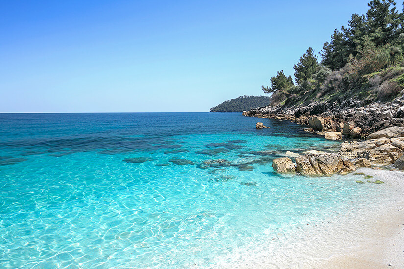 Eine kleine Bucht mit hellem Kies- oder Sandstrand geht in kristallklares, türkisfarbenes Wasser über. Felsen und bewaldete Hänge säumen die Küste und verleihen der Landschaft einen natürlichen, unberührten Charakter. Thassos Strand
