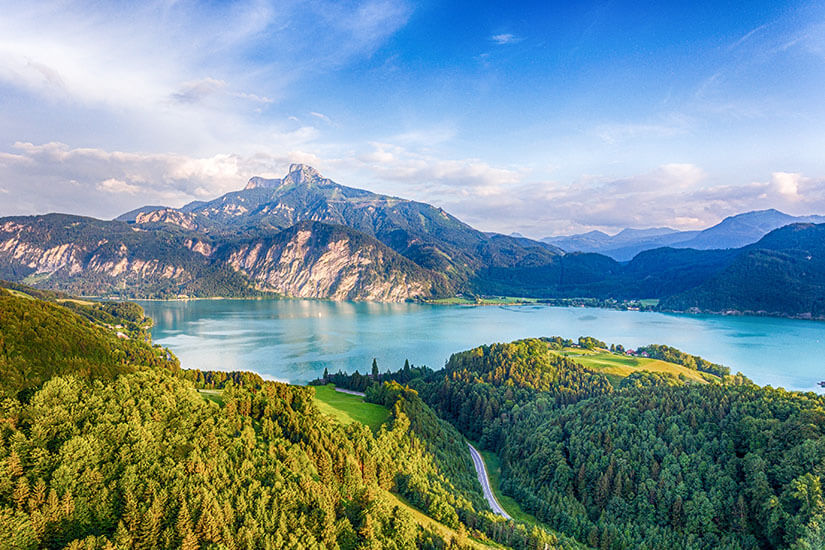 Ein großer See liegt eingebettet zwischen bewaldeten Hügeln und markanten Bergen, deren steile Felswände bis ans Ufer reichen. Im Vordergrund schlängelt sich eine Straße durch grüne Wiesen und dichte Wälder. Mondsee See