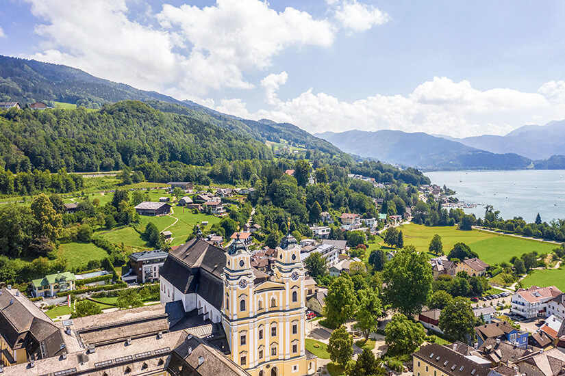 Eine Ortschaft mit einer markanten Kirche liegt eingebettet zwischen grünen Hügeln und einem großen See. Im Hintergrund erheben sich bewaldete Berge unter einem teilweise bewölkten Himmel. Mondsee Gemeinde