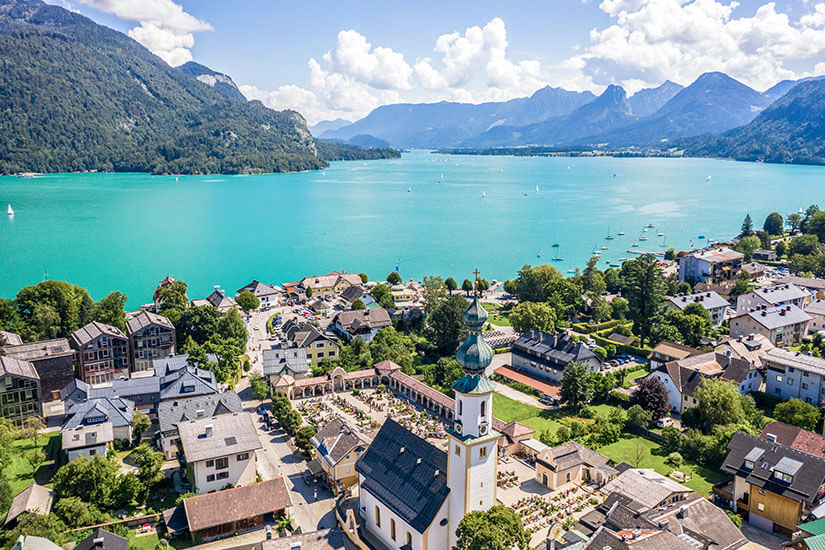 Eine Ortschaft mit einer Kirche und ihrem markanten Zwiebelturm liegt direkt am Ufer eines türkisfarbenen Sees. Zahlreiche Boote sind auf dem Wasser zu erkennen, während bewaldete Berge die Landschaft im Hintergrund umrahmen. Oesterreich St Gilgen