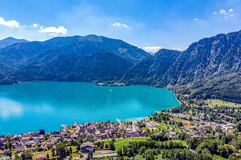 Ein Ort mit dicht stehenden Häusern liegt direkt am Ufer eines türkisfarbenen Sees und wird von bewaldeten Bergen umgeben. Entlang der Küste sind ein kleiner Hafen, Grünflächen und Freizeitanlagen zu erkennen. Attersee Unterach