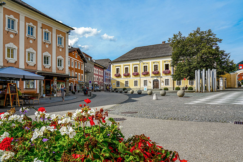 Ein gepflasterter Marktplatz wird von farbenfrohen historischen Gebäuden mit Cafés und Geschäften umrahmt. Im Vordergrund sorgen blühende Blumen für einen lebendigen Farbakzent, während auf dem Platz moderne Gestaltungselemente und wenige Passanten zu sehen sind. Mondsee Marktplatz