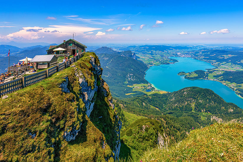 Auf einem steilen Berggipfel befindet sich eine Hütte mit Aussichtsterrasse und Geländern, von der aus Besucher die Landschaft genießen können. Tief darunter erstreckt sich ein großer türkisblauer See, umgeben von bewaldeten Hügeln, Bergen und kleinen Ortschaften. Oesterreich Schafberg