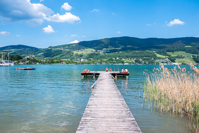 Ein langer Holzsteg führt in einen ruhigen See, an dessen Ende mehrere Menschen sitzen oder sich sonnen. Im Hintergrund sind grüne Hügel mit verstreuten Häusern zu sehen, während Schilf am Ufer wächst. Mondsee Baden