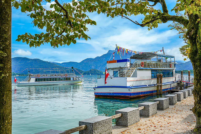 Zwei Ausflugsschiffe liegen auf einem ruhigen See, eingerahmt von Bäumen am Ufer und einer befestigten Promenade. Im Hintergrund erheben sich bewaldete Berge unter einem leicht bewölkten Himmel. Oesterreich Schifffahrt