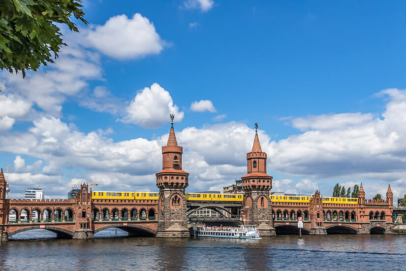 Die markante Oberbaumbrücke aus rotem Backstein verbindet Kreuzberg und Friedrichshain über die Spree. Auf ihr rollt die charakteristische gelbe U-Bahn der Linie U1, darunter schippert ein Ausflugsboot vorbei. Städtereisen Bahn Berlin