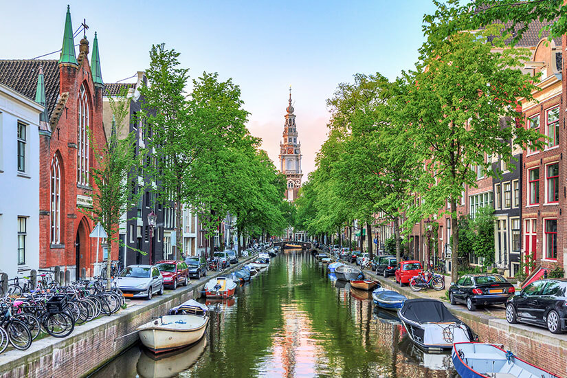 Blick durch eine typische Amsterdamer Gracht, gesäumt von Backsteinhäusern und dichten Baumkronen. Zwischen Fahrrädern und vertäuten Booten spiegelt sich am Ende der Turm der Zuiderkerk im ruhigen Wasser. Städtereisen Bahn Amsterdam