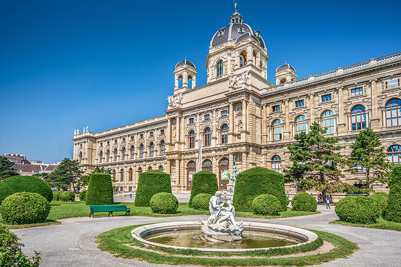 Der prachtvolle Bau des Naturhistorischen Museums im Wiener Museumsquartier mit seiner charakteristischen Kuppel und aufwändig verzierter Fassade. Im Vordergrund ein gepflegter Park mit Brunnen und Skulpturen. Städtereisen Bahn Wien