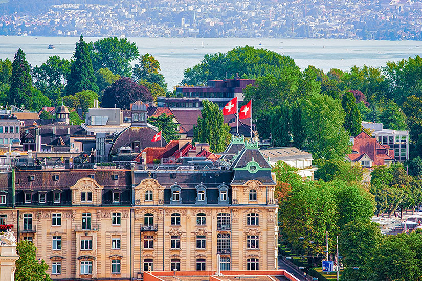 Über den Dächern Zürichs öffnet sich der Blick auf den weiten Zürichsee, im Vordergrund wehen zwei Schweizer Fahnen im Sommerwind. Elegante Fassaden und viel Grün prägen das Motiv. Städtereisen Bahn Zürich
