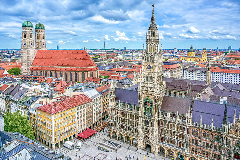 Von oben blickt man auf den Marienplatz mit dem neogotischen Neuen Rathaus samt Glockenspielturm. Im Hintergrund erheben sich die beiden charakteristischen Zwiebeltürme der Frauenkirche über den Dächern Münchens. Städtereisen Bahn München