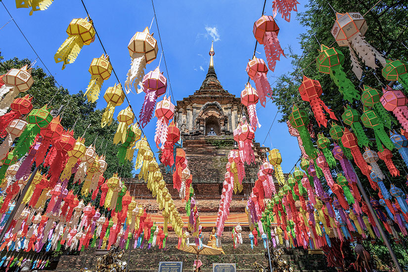 Ein historischer Tempel mit einer hohen, spitz zulaufenden Pagode wird von zahlreichen bunten Laternen geschmückt, die über dem Weg aufgehängt sind. Die farbenfrohen Dekorationen bilden einen auffälligen Kontrast zum blauen Himmel und der alten Backsteinarchitektur. Workation Thailand