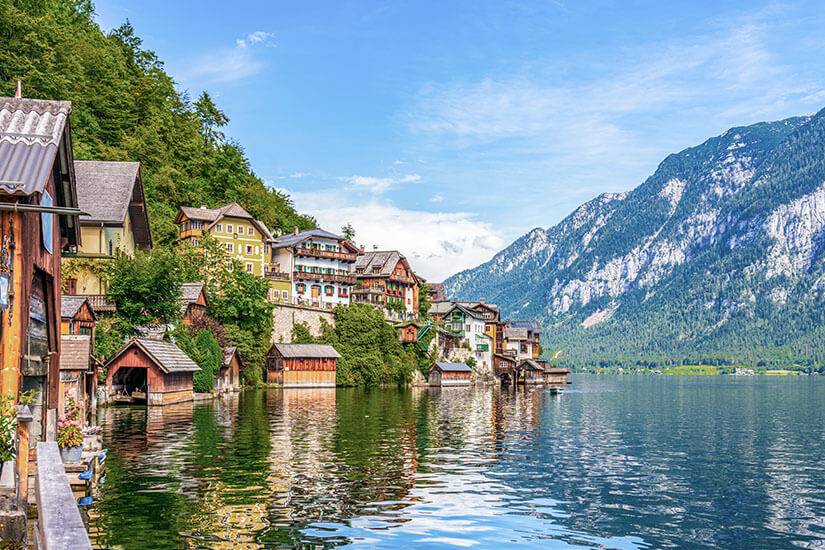 Ein malerisches Dorf mit traditionellen Häusern schmiegt sich an den Hang eines Berges direkt am Ufer eines ruhigen Sees. Mehrere kleine Bootshäuser stehen unmittelbar am Wasser, während sich die umliegenden Berge in der glatten Oberfläche spiegeln. Workation Oesterreich