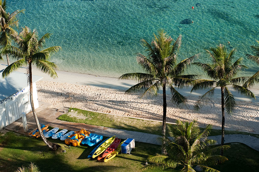 Palmen säumen einen hellen Strand, an dem Kajaks und kleine Boote bereitliegen. Das Wasser ist klar und ruhig, weshalb die Szene besonders familienfreundlich wirkt. Der Blick von oben vermittelt gut, wie nah Erholung, Wassersport und einfache Strandtage auf Guam beieinanderliegen. Guam Mikronesien Strand