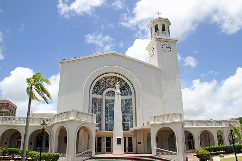 Die weiße Fassade der Kathedrale steht hell vor blauem Himmel und tropischem Grün. Der Bau wirkt schlicht, aber durch Turm, Rundbogen und großes Glasfenster sehr markant. Guam Mikronesien Kirche