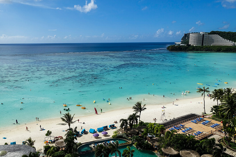 Die Tumon Bay wirkt hier wie ein klassischer Urlaubsstrand: helles Ufer, ruhiges Lagunenwasser und viele Badende im flachen Meer. Am Rand stehen Resorts, Liegen und Palmen, wodurch der touristische Charakter dieses Küstenabschnitts deutlich wird. Guam Mikronesien Tumon Bay Beach