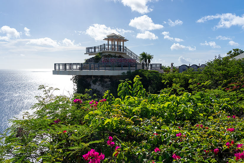 Die Aussichtsplattform ragt über eine grüne Klippe hinaus und öffnet den Blick auf das weite Meer. Pinke Blüten und dichter Bewuchs rahmen den Vordergrund, während der Pazifik im Hintergrund fast grenzenlos wirkt. Guam Mikronesien Two Lovers Point