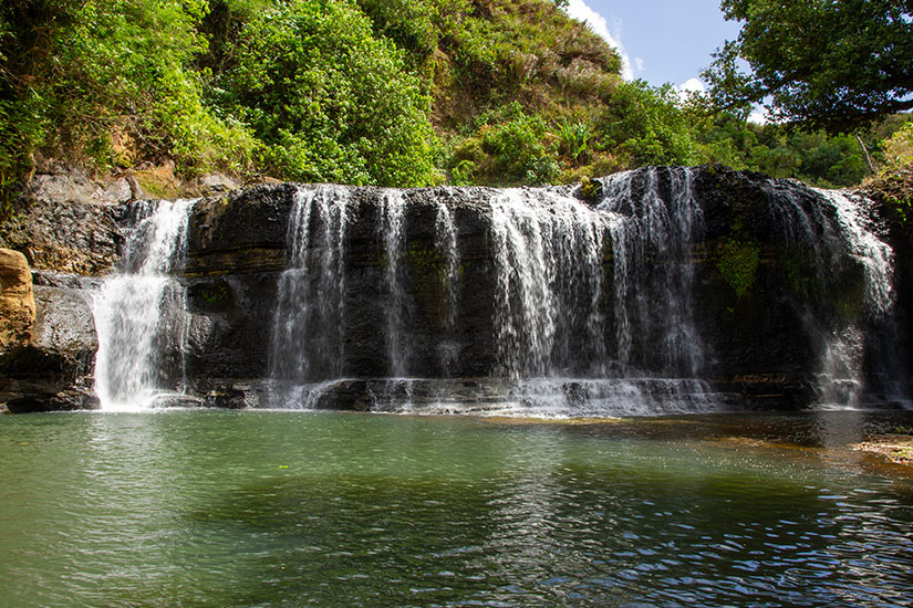 Breite Wasservorhänge fallen über dunkle Felsen in ein ruhiges Becken. Rundherum wächst dichtes Grün, wodurch der Ort abgeschieden und naturbelassen wirkt. Guam Mikronesien Wasserfall