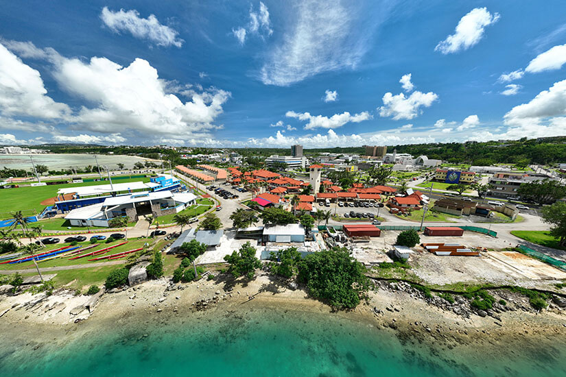 Der Küstenbereich wirkt lebendig, aber nicht überfüllt: Häuser, Straßen, Sportflächen und Wasser liegen nah beieinander. Im Hintergrund ziehen sich grüne Hügel und Hotelanlagen entlang der Bucht. Guam Mikronesien Sengsong