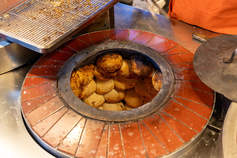 Mehrere runde Fladenbrote backen in einem tiefen, heißen Ofen und haben bereits eine goldbraune Kruste. Die Backstation wirkt wie Teil einer offenen Küche oder eines Marktstands, an dem die Brote frisch zubereitet werden. Taiwan Essen Traditionen