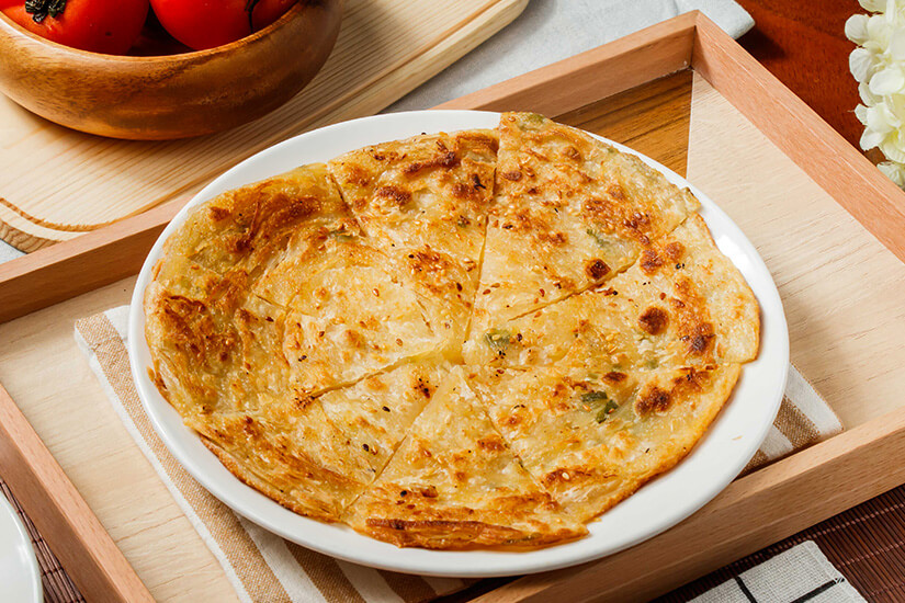 Auf einem weißen Teller liegt ein goldbraun gebackener, in Stücke geschnittener Fladen oder Pfannkuchen. Er wird auf einem Holztablett serviert, daneben sind unter anderem eine Schüssel mit Tomaten zu sehen. Taiwan Essen Pfannkuchen