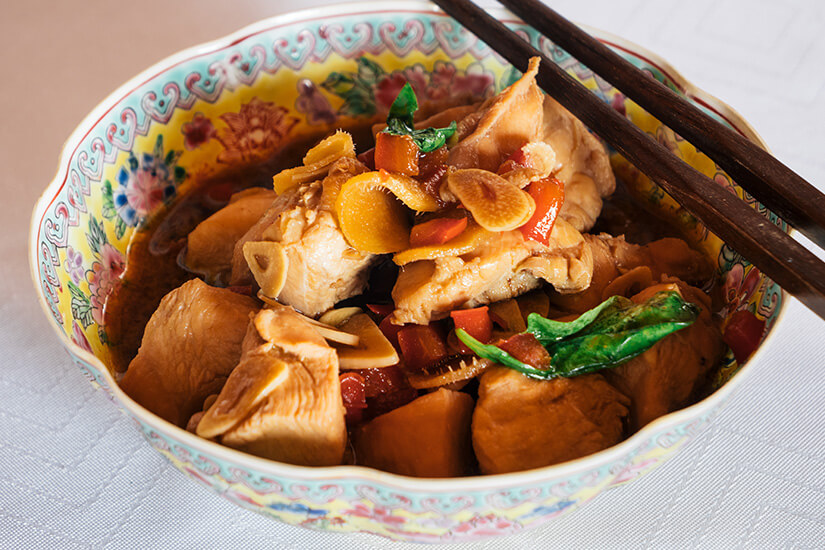 In einer dekorativen Schüssel wird ein geschmortes Gericht mit Fleischstücken, Gemüse und Kräutern in einer dunklen Sauce serviert. Daneben liegen Essstäbchen, die auf eine asiatische Tischkultur hindeuten. Taiwan San Bei Ji
