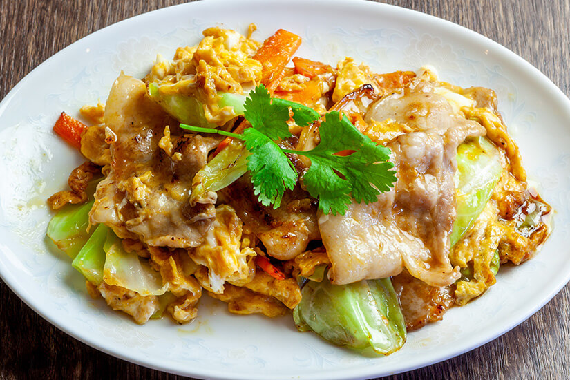 Auf einem weißen Teller liegt ein warmes Pfannengericht mit Fleisch, Ei und verschiedenen Gemüsesorten wie Kohl und Karotten. Ein frischer Kräuterzweig dient als Garnitur und setzt einen grünen Farbakzent. Taiwan Hakka Pfanne