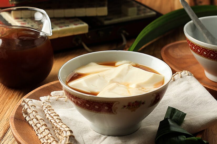 In einer Schüssel befindet sich ein cremiger, heller Nachtisch, der mit einer braunen Sirup- oder Zuckersauce übergossen ist. Daneben stehen ein Kännchen mit weiterer Sauce sowie Geschirr auf einem hölzernen Tisch. Taiwan Essen Nachtisch