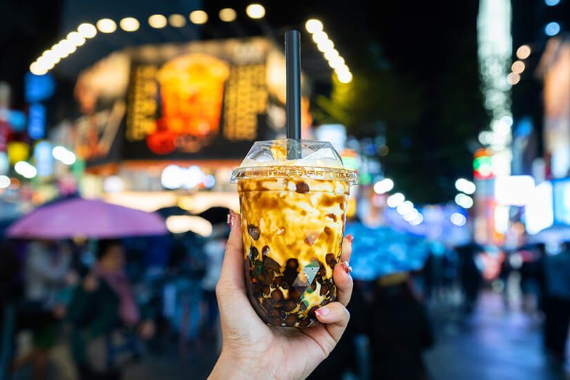Eine Hand hält einen Becher mit einem Getränk, das schwarze Tapiokaperlen und eine cremige Schicht enthält, durch die ein breiter Trinkhalm steckt. Im Hintergrund sind unscharfe Lichter, Menschen und eine belebte Straße bei Nacht zu erkennen. Taiwan Bubble Tea