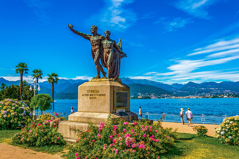 Im Vordergrund steht ein Denkmal mit zwei Bronzefiguren auf einem steinernen Sockel, umgeben von blühenden Rosen und gepflegten Grünanlagen. Dahinter verlaufen eine Uferpromenade und ein großer See, vor dessen Kulisse sich Berge unter einem klaren Himmel erheben. Einige Menschen spazieren entspannt entlang des Wassers. Stresa Promenade