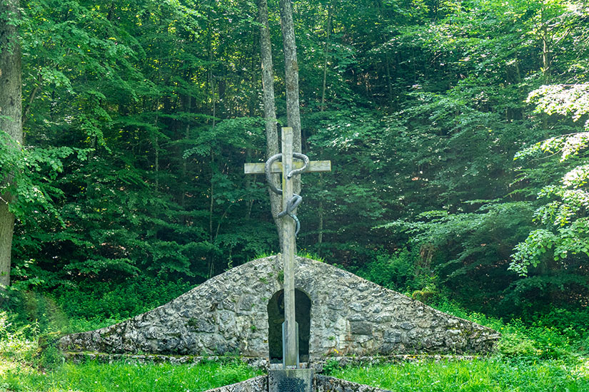 Ein steinernes Kreuz steht im grünen Wald. Pfronten Allgaeu Kalvarienberg