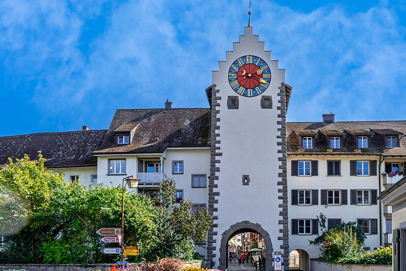 Die straßenseitige Ansicht des historischen Untertors in Stein am Rhein. Der weiße, hohe Torturm besitzt an den Ecken markante, graue Ecksteine und schließt nach oben mit einem steinernen Treppengiebel ab. Im oberen Bereich des Turms prallt eine große, runde Turmuhr mit einem rot-blauen Ziffernblatt und goldenen römischen Ziffern und Zeigern ins Auge. Durch den großen, rundbogigen Torbogen im Erdgeschoss ist der Blick auf Passanten und die historische Altstadt dahinter freigegeben, während links und rechts weiße Wohngebäude mit dunklen Dächern an den Turm anschließen. Stein am Rhein Schweiz Untertor