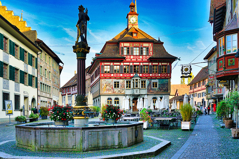 Ein zentraler Blick auf den Marktplatz in Stein am Rhein mit einem historischen Marktbrunnen im Vordergrund. Der steinerne Brunnen besitzt ein achteckiges Becken und eine kunstvolle Mittelsäule, die von einer Ritterstatue gekrönt wird und mit bunten Blumenkästen geschmückt ist. Im Hintergrund steht das markante Rathaus, ein prachtvolles Fachwerkgebäude mit kunstvollen Fassadenmalereien, roten Holzbalken und einem kleinen Uhrturm auf dem Dach, umgeben von gepflasterten Gassen und Außengastronomie. Stein am Rhein Schweiz Rathaus