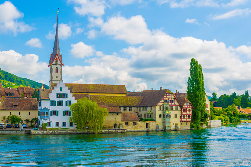 Blick von der gegenüberliegenden Flussseite auf das ehemalige Kloster St. Georgen direkt am Ufer des tiefblauen Rheins. Die historische Gebäudeanlage zeigt links ein weißes Haus mit grünen Fensterläden und Treppengiebel, hinter dem der markante, spitz zulaufende Kirchturm mit roter Umrandung aufragt. Rechts schließen sich weitere klösterliche Bauten mit hellem Fachwerk und Erkern an, umgeben von grünen Bäumen unter einem leicht bewölkten, blauen Himmel. Stein am Rhein Schweiz St Georgen