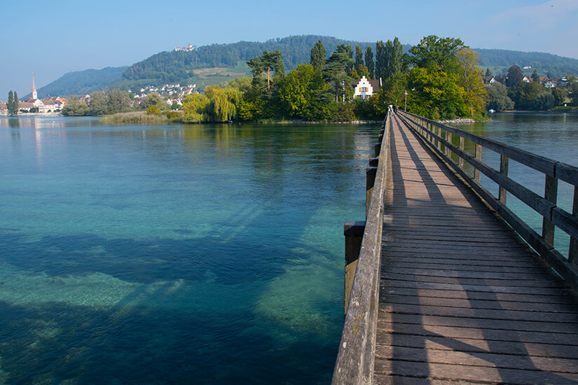 Ein langer Holzsteg mit einfachen Holzgeländern führt von der rechten unteren Bildecke geradeaus über das ruhige, klare Flusswasser, das im Vordergrund helle Türkistöne aufweist und den Blick auf den Grund freigibt. Am Ende des Stegs befindet sich eine grüne Flussinsel mit Bäumen und einem weißen Wohnhaus mit Treppengiebel. Im linken Hintergrund erstreckt sich die Silhouette von Stein am Rhein mit einem Kirchturm, während im fernen Hintergrund eine bewaldete Hügelkette mit einer Burg auf der Bergkuppe unter einem leicht dunstigen, blauen Himmel zu sehen ist. Stein am Rhein Schweiz Panoramaweg