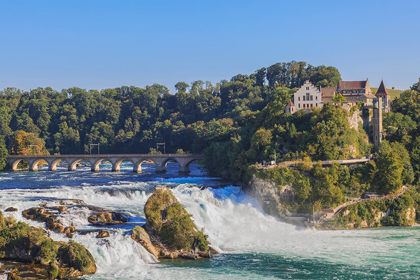 Ein weiter Blick auf die tosenden Wassermassen des Rheinfalls, die im Vordergrund weiß schäumend über Felsen stürzen. Mittendrin ragt ein großer, mit Grün bewachsener Felsen empor. Im Hintergrund überspannt eine historische Steinbogenbrücke den ruhigeren Flussabschnitt vor einer dichten Kulisse aus grünen Bäumen. Auf einem steilen Felsvorsprung auf der rechten Seite thront das Schloss Laufen mit seinen hellen Mauern und runden Turmelementen unter einem klaren, blauen Himmel. Stein am Rhein Schweiz Rheinfall