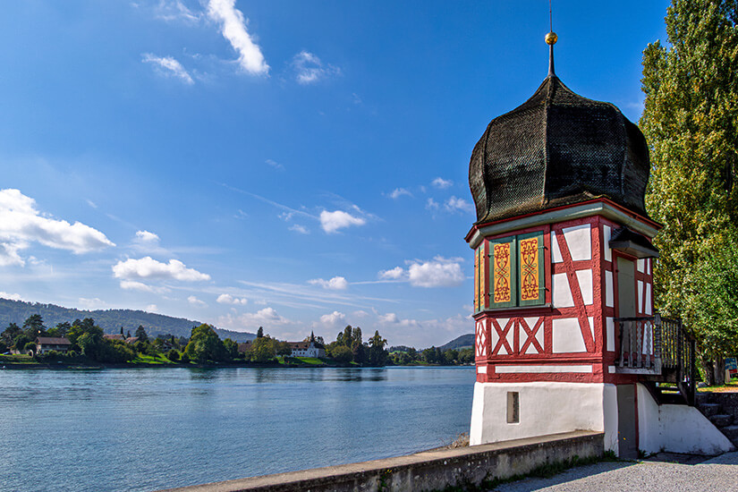 Ein kleiner, charmanter Pavillon in Fachwerkbauweise steht direkt am befestigten Flussufer des ruhig dahinfließenden Rheins. Das Häuschen besticht durch seine rot-weißen Balken, kunstvoll verzierte, gelb-rote Fensterläden und ein markantes, dunkles Zwiebeldach mit einer kleinen goldenen Spitze. Auf der gegenüberliegenden Flussseite erstreckt sich eine grüne Landschaft mit Bäumen, vereinzelten Wohnhäusern und einer kleinen Kirche vor einer bewaldeten Hügelkette unter einem strahlend blauen Himmel mit lockeren Wolken. Stein am Rhein Ausblick auf den Rhein