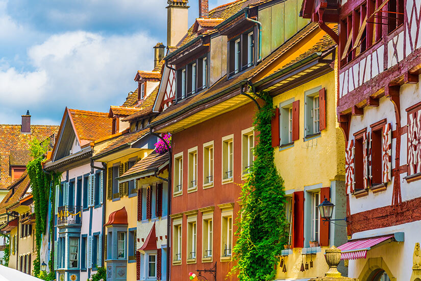 Eine dichte Reihe von historischen, mehrstöckigen Hausfronten in einer gepflasterten Altstadtgasse. Die Fassaden zeigen eine bunte Vielfalt an Farben von kräftigem Rotbraun über helles Gelb bis hin zu zartem Blau und Weiß. Viele Gebäude besitzen traditionelles Fachwerk, dekorative Dachgauben und hölzerne Fensterläden, während im Vordergrund eine Hauswand üppig mit grünen Kletterpflanzen bewachsen ist und ein rot-weiß gestreiftes Vordach ins Auge fällt. Stein am Rhein Schweiz Hausfronten