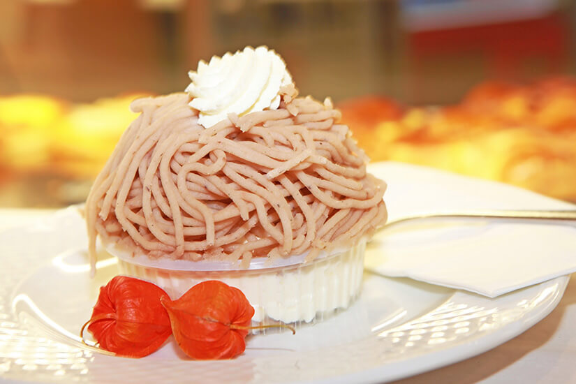 Ein Nahaufnahme einer traditionellen Schweizer Vermicelles-Süßspeise, die auf einem weißen Teller serviert wird. Das Dessert besteht aus einem runden Baiser-Boden, auf dem das hellbraune Kastanienpüree in langen, wurmartigen Fäden zu einer Haube aufgespritzt und mit einem Klecks Schlagsahne garniert ist. Vor dem Dessert liegen zwei orangefarbene Lampionblumen zur Dekoration auf dem Teller, während der Hintergrund unscharf gehalten ist. Stein am Rhein Schweiz Vermicelles