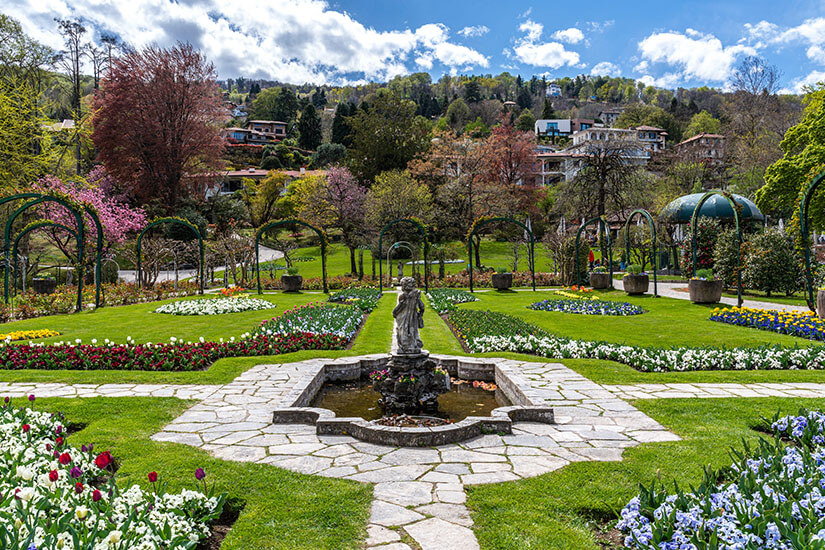 Ein symmetrisch angelegter Park mit gepflegten Blumenbeeten, Wegen und einer kleinen Brunnenfigur erstreckt sich bis zu einem bewaldeten Hang mit Häusern. Im Hintergrund sind Pavillons, Bäume und blühende Pflanzen zu sehen. Stresa Pallavicino