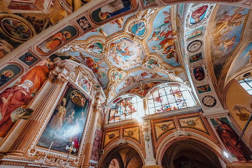 Eine reich verzierte Kirchenhalle mit kunstvollen Deckenfresken, religiösen Gemälden und dekorativen Stuckelementen ist zu sehen. Großflächige Fenster lassen Sonnenlicht in den Innenraum fallen und beleuchten die farbenprächtige Architektur. Piemont Kloster