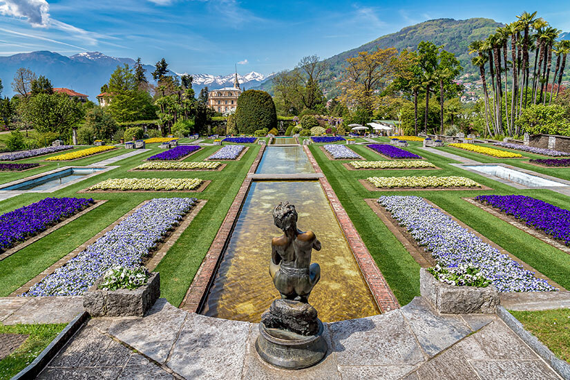 Zu erkennen ist ein prachtvoll angelegter Barockgarten mit symmetrischen Blumenbeeten, langen Wasserbecken und einer Statue im Vordergrund. Im Hintergrund stehen elegante Gebäude, hohe Palmen sowie bewaldete Berge mit teils schneebedeckten Gipfeln unter einem klaren blauen Himmel. Piemont Villa Taranto