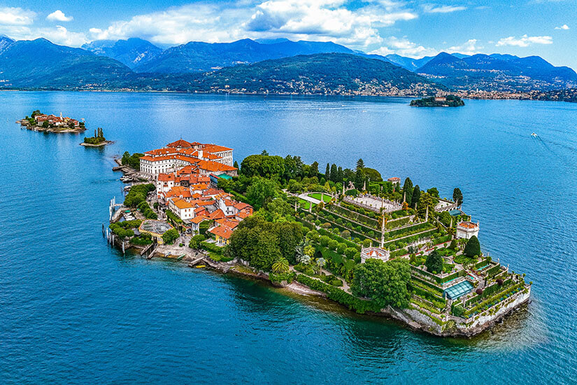 Zu erkennen ist eine dicht bebaute Insel mit historischen Gebäuden, terrassenförmig angelegten Gärten und üppiger Vegetation, umgeben von einem großen See. Im Hintergrund liegen weitere kleine Inseln sowie eine Berglandschaft unter einem teilweise bewölkten Himmel. Stresa Inseln