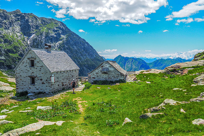 Zu erkennen ist eine alpine Landschaft mit zwei steinernen Berghütten auf einer grünen Wiese, umgeben von schroffen Felsen und hohen Bergen. Vor einer der Hütten steht eine Person, während am Horizont weitere Bergketten unter einem Himmel mit großen Wolken zu sehen sind. Stresa Wandern