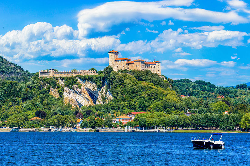 Zu erkennen ist eine mittelalterliche Burg auf einem bewaldeten Hügel über einem See. Im Vordergrund fährt ein kleines Motorboot über das Wasser, während im Hintergrund dichte Wälder und ein Himmel mit markanten Wolkenformationen zu sehen sind. Lago Maggiore Rocca di Angera